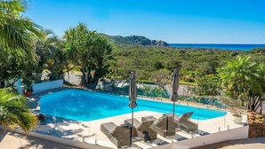 A swimming pool with lounge chairs and umbrellas, surrounded by lush greenery and a view of the ocean. - Byron Pacific Vista - Family Fun with Pool (Byron)