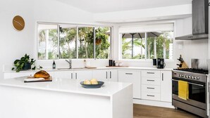 A modern kitchen with white cabinetry, a central island, and large windows offering a view of greenery. - Byron Pacific Vista - Family Fun with Pool (Byron)