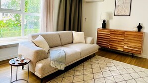 A living room with a beige sofa, a wooden sideboard, a patterned rug, and a window with curtains. - Villa Inn Sintra | Private Garden Retreat (Sintra)