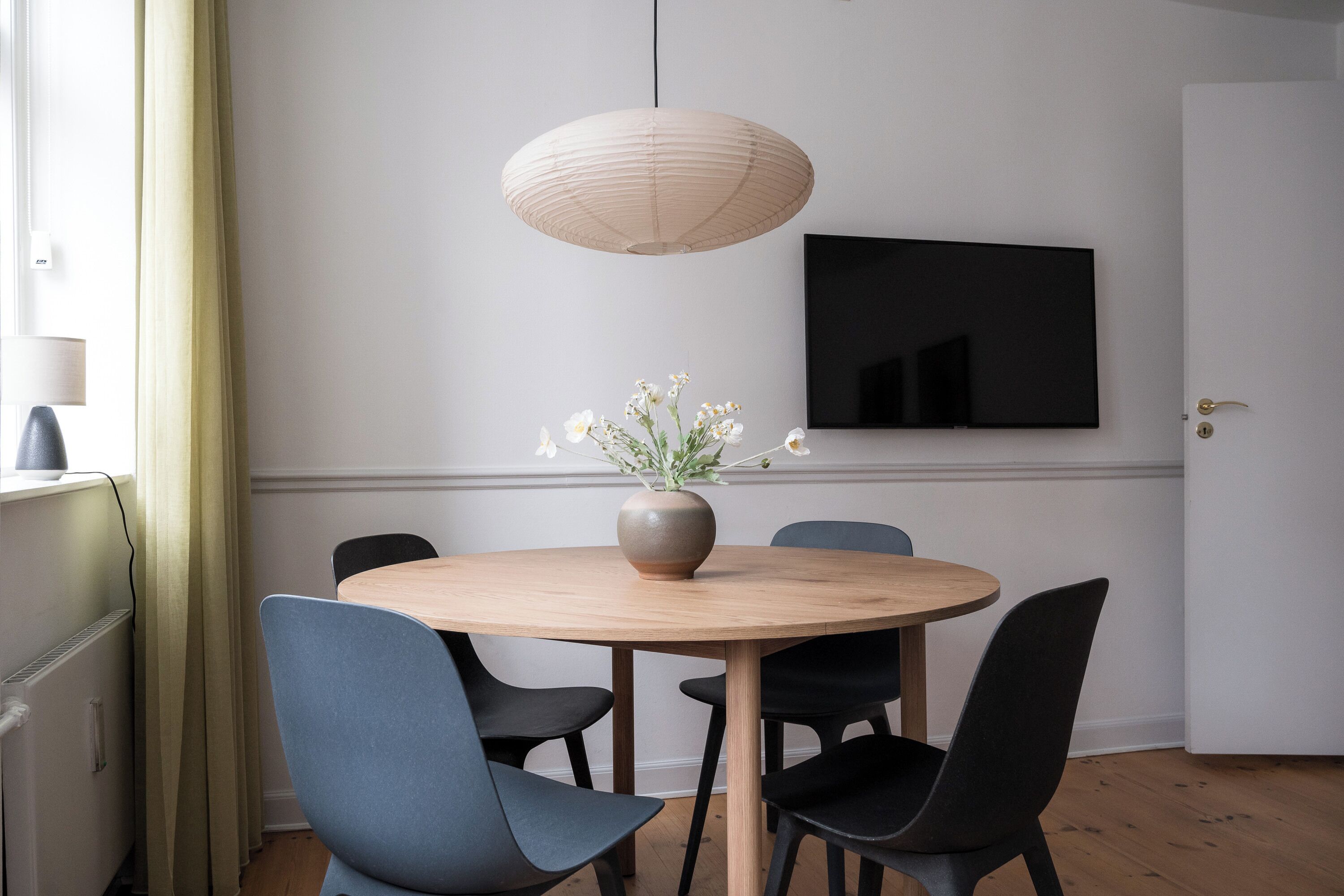 Un comedor con una mesa redonda de madera, sillas negras, un florero con flores, una lámpara colgante y un televisor de pantalla plana en la pared.
