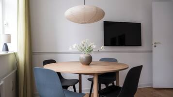 Un comedor con una mesa redonda de madera, sillas negras, un florero con flores, una lámpara colgante y un televisor de pantalla plana en la pared.