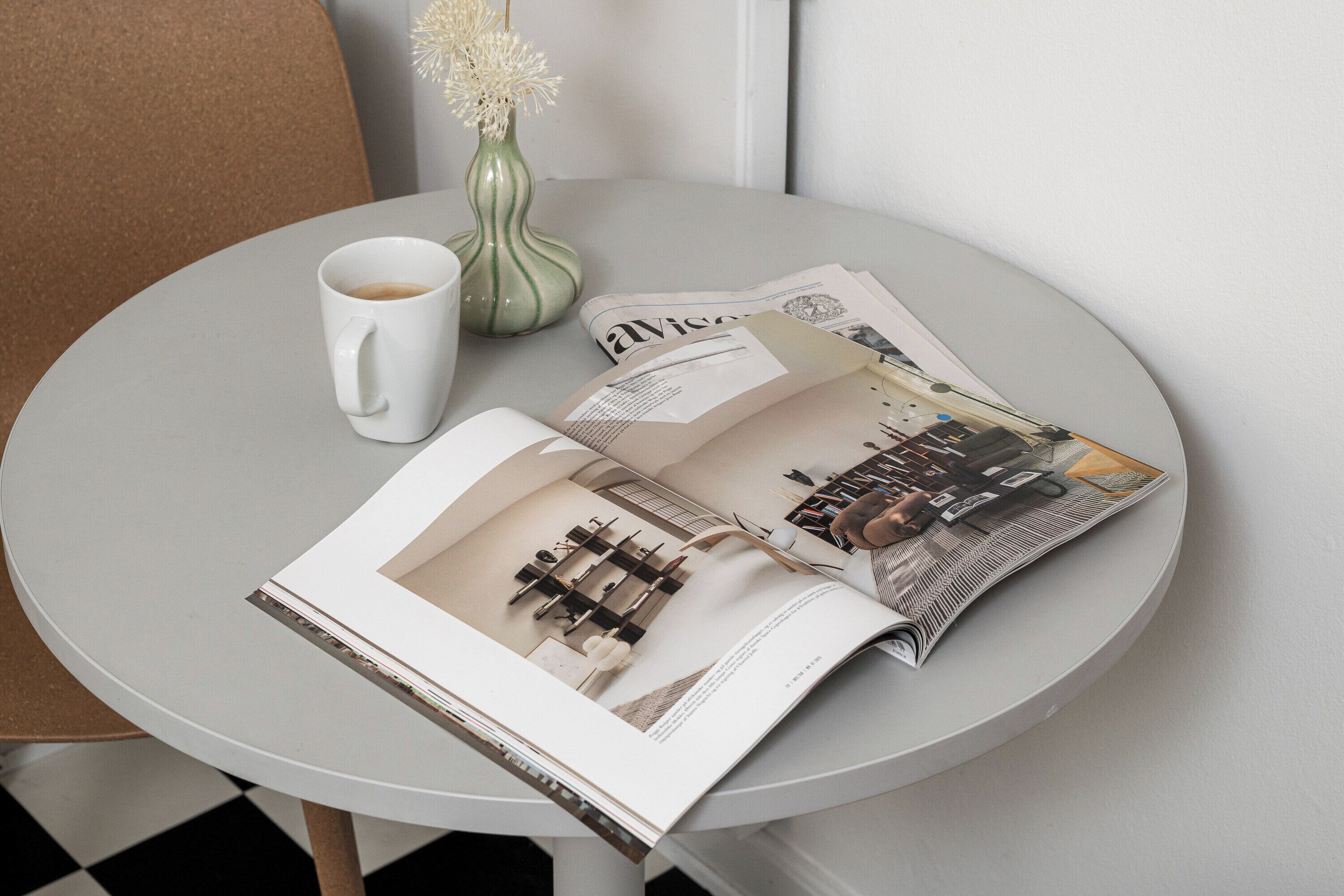 A magazine on a round table with a white mug and a vase of flowers.