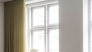 Una habitación con un sillón blanco, una mesita de noche con una lámpara y una ventana con cortina verde.