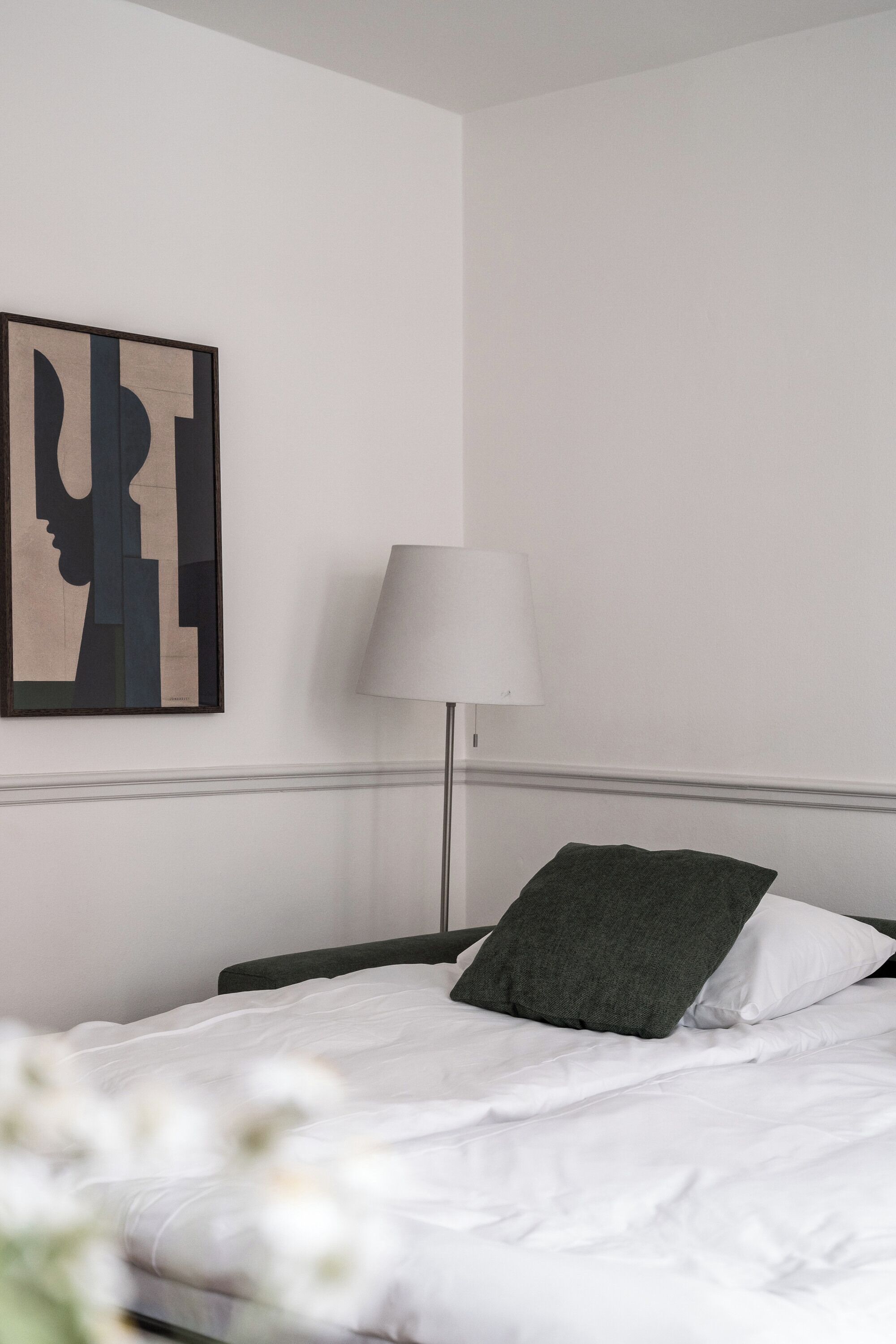 Un lit bien fait, avec du linge de lit blanc et un oreiller foncé, une lampe moderne posée à côté, et une peinture abstraite minimaliste accrochée au mur.