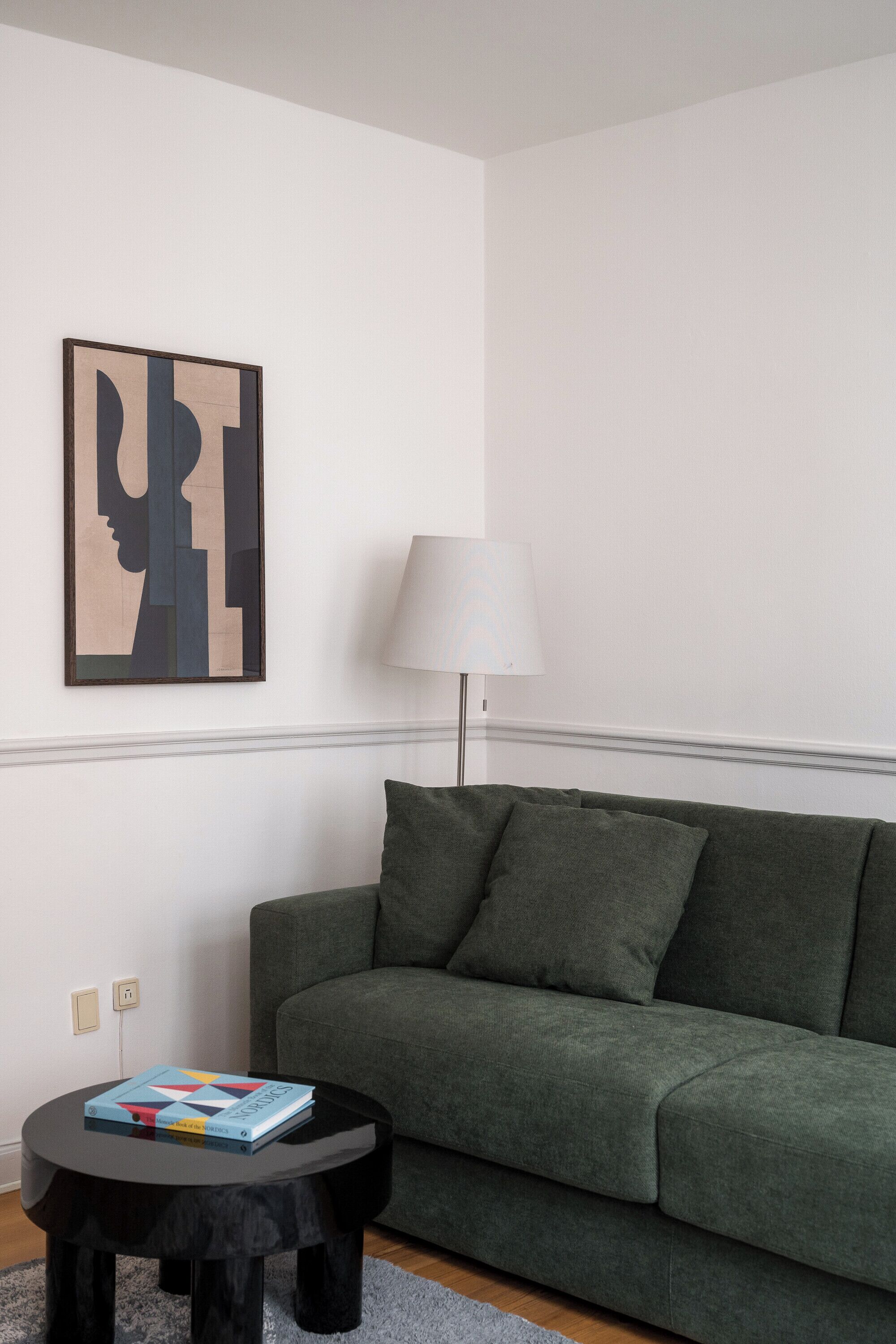 A modern living room with a green sofa, a black coffee table with a book, a lamp, and a wall-mounted abstract art piece.