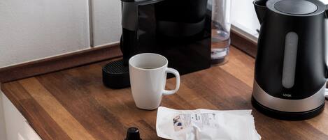 A kitchen counter with a coffee maker, a mug, a kettle, and a packet of coffee pods.