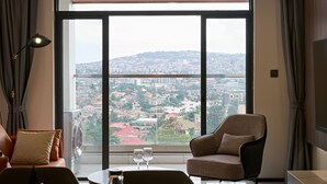 A modern living room with a large window offering a city view, a sofa, a chair, and a table with glasses. - 3-Bedroom apartment at Oasis Park. (Kigali)