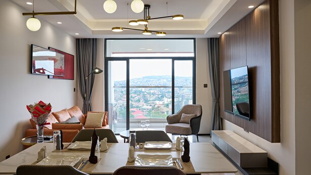 A modern living room with a dining area, large windows with a city view, and a mounted TV. - 3-Bedroom apartment at Oasis Park. (Kigali)