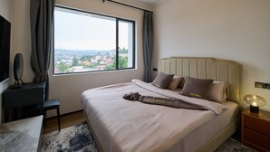 A hotel room with a large window, a bed with beige bedding, a bedside table with a lamp, and a desk with a chair.