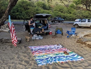 Un camping con un vehículo, sillas de camping y una manta sobre la arena.