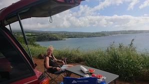 Une personne est assise à une table devant une pastèque, une voiture, et un paysage pittoresque comprenant un lac et une forêt.