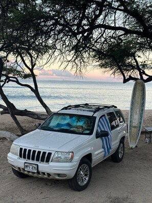 Un SUV blanc aparcat a una platja sorrenca, amb una taula de surf recolzada contra ell.