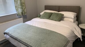 Een keurig opgemaakt bed met witte lakens en twee groene kussens, een nachtkastje met een lamp, en een raam met zonwering en een gordijn met bladmotief.