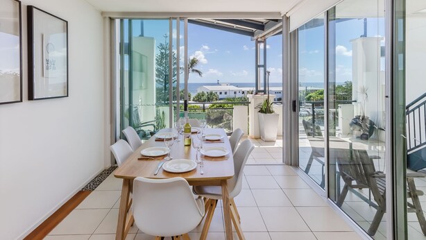 Un coin repas avec une table dressée pour quatre personnes, de grandes fenêtres offrant une vue sur la mer et un balcon aménagé avec du mobilier d’extérieur.