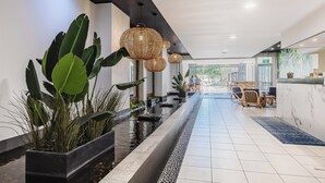 Un hall d’hôtel moderne avec un long comptoir d’accueil, des plantes en pot, un élément décoratif avec de l’eau et des suspensions lumineuses en osier.