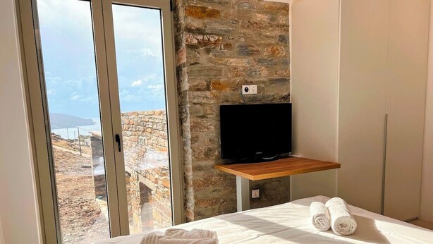 A bedroom with a bed, a television on a wooden shelf, a stone wall, and a view through a glass door. - Tzia Stone Modern Studio with Sea View (Kea)