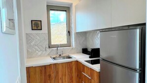 A modern kitchen with white cabinets, a stainless steel refrigerator, and a window providing natural light. - Tzia Stone Modern Studio with Sea View (Kea)