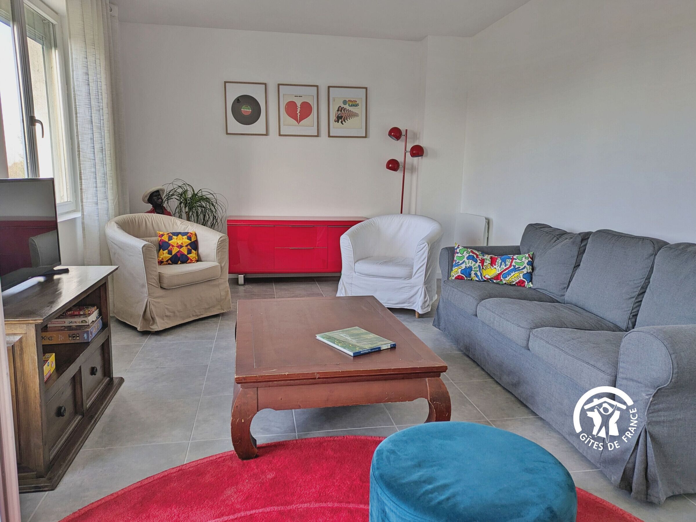 A living room with a sofa, armchair, coffee table, and a red cabinet. There are three framed pictures on the wall.