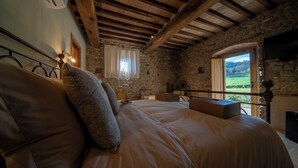 Een slaapkamer met een groot bed, stenen muren, houten balken, een raam met gordijnen en uitzicht op groen.