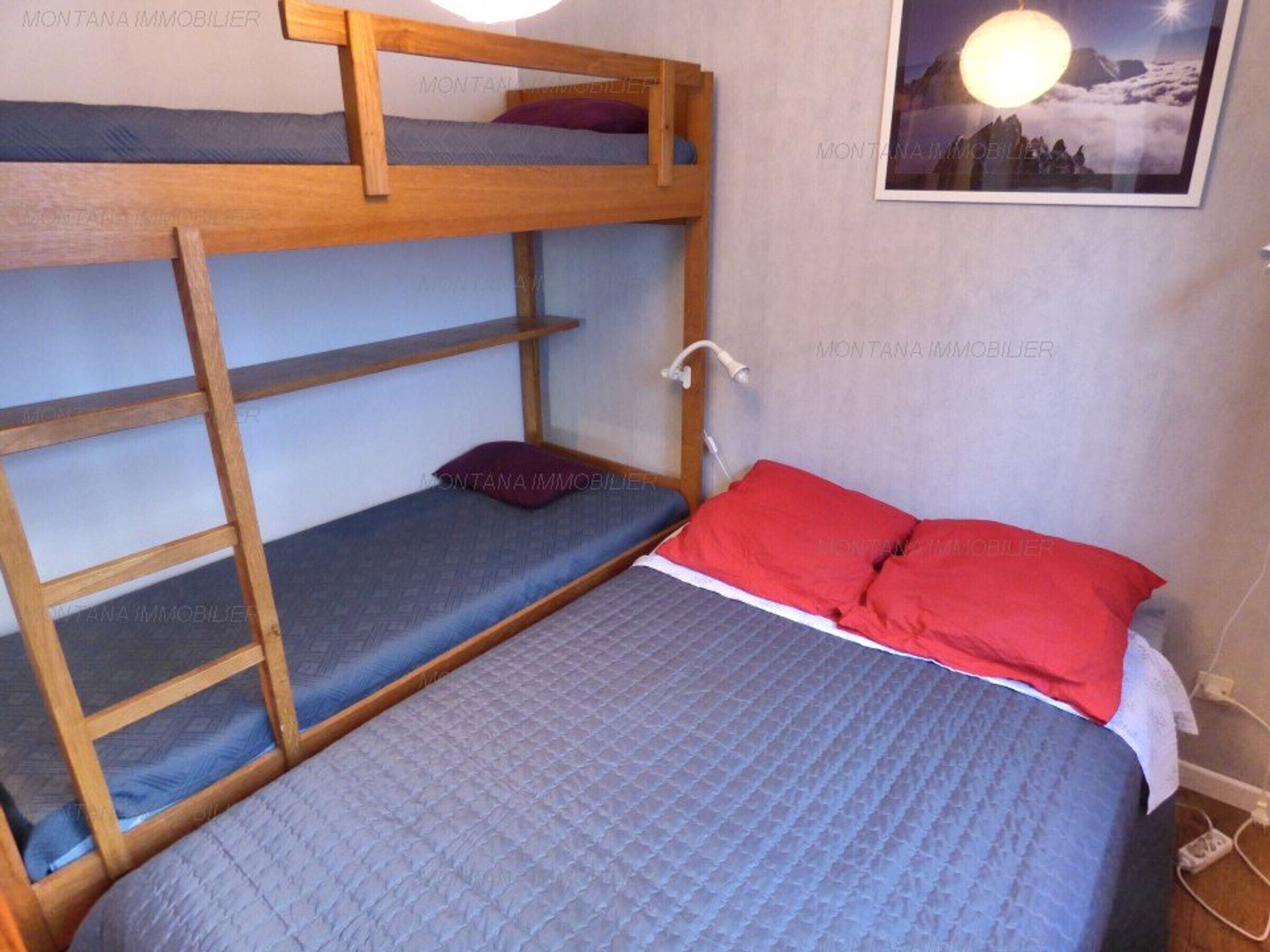 A bunk bed room with a wooden ladder, a single bed with red pillows, and a wall-mounted light.