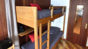 Une chambre avec des lits superposés, des murs en bois, une porte dotée d’une fenêtre en verre dépoli et une petite table avec un haut-parleur.