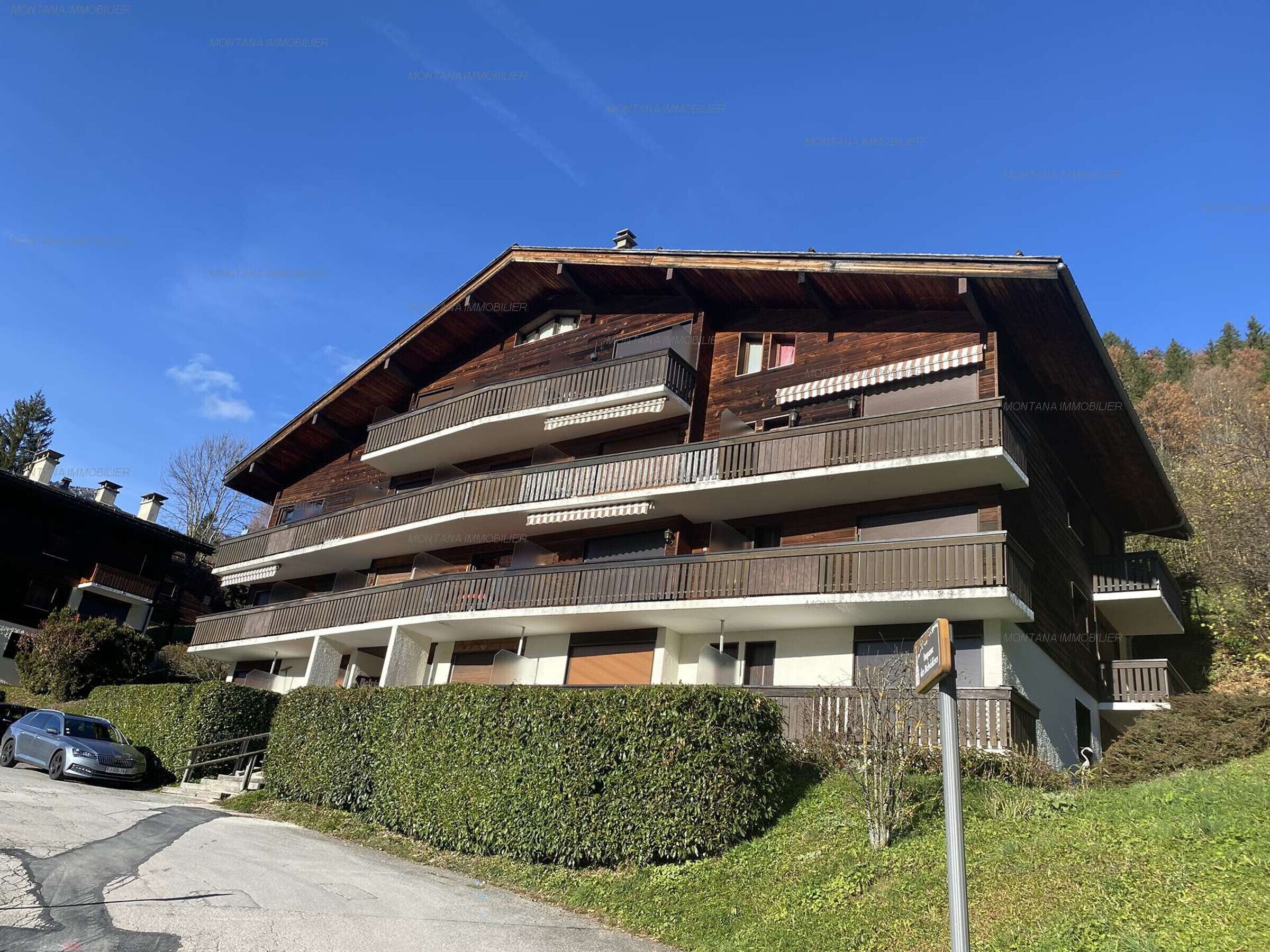 A multi-story wooden building with balconies, a paved driveway, and a well-maintained hedge.