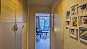 Ein Flur, der zu einem Balkon mit Blick auf schneebedeckte Bäume führt. An der Wand hängen Bilder in Rahmen und eine Tür mit Griff.