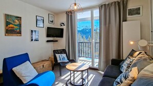 Ein modernes Wohnzimmer mit einem blauen Sofa, einem grauen Sofa, einem kleinen runden Tisch und einem an der Wand montierten Fernseher. An den Wänden befinden sich gerahmte Bilder, ein Fenster mit Blick auf Bäume und ein Vorhang.