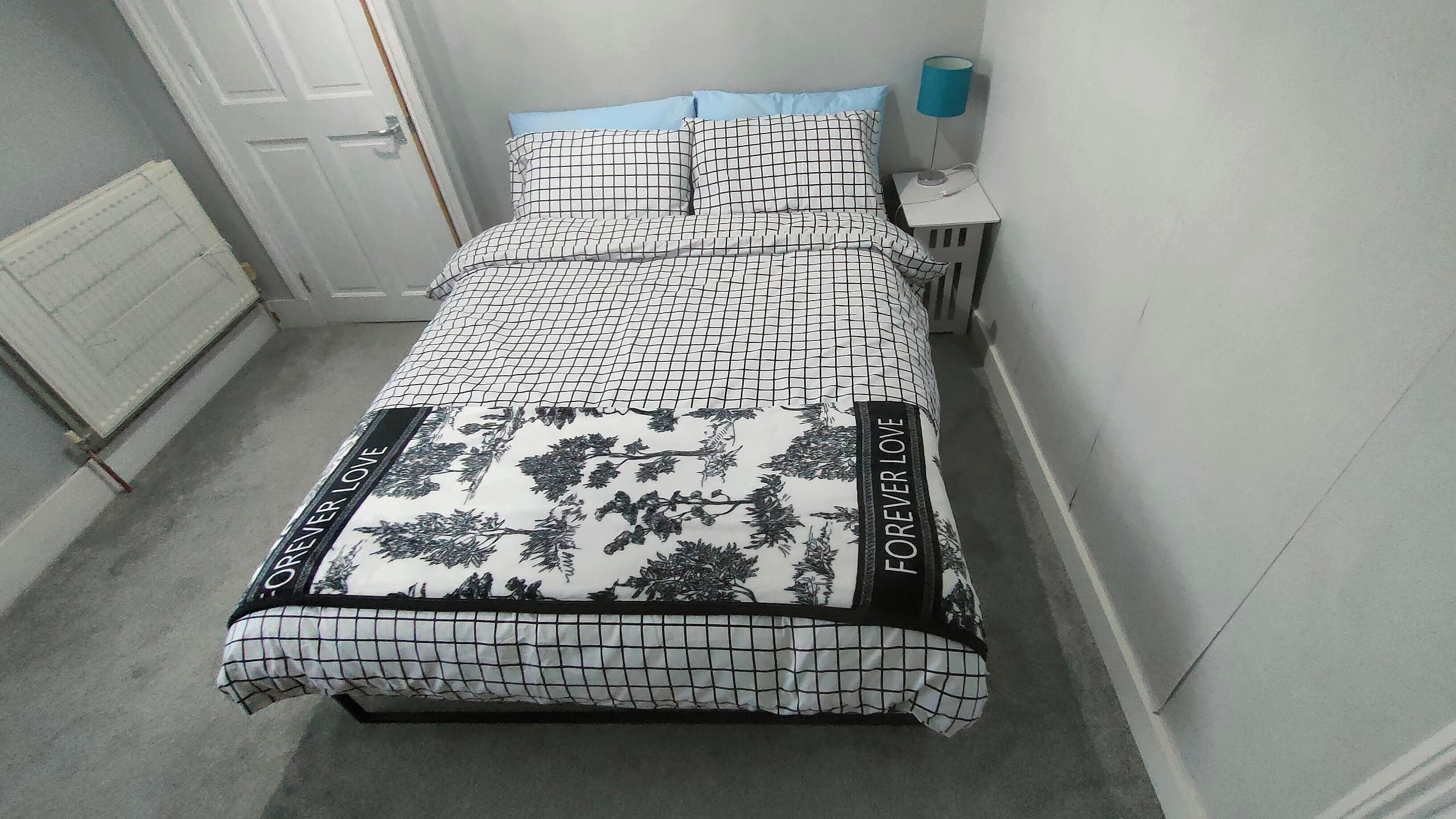 Een bed met een geruit patroon en bloemenmotief, een nachtkastje met een lamp, en een deur op de achtergrond.