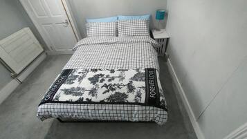 Een bed met een geruit patroon en bloemenmotief, een nachtkastje met een lamp, en een deur op de achtergrond.