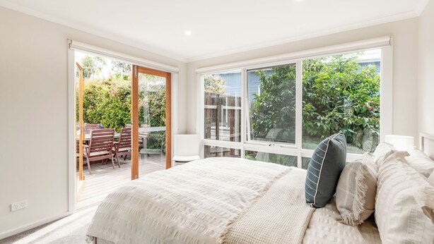 A bedroom with a large bed, a sliding glass door leading to a patio, and a view of greenery outside. - Geelong Road (Barwon Heads)