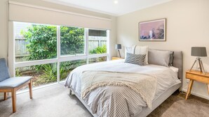 A bedroom with a large window, a bed with striped bedding, a chair, and a bedside table with a lamp. - Geelong Road (Barwon Heads)
