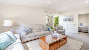 A modern living room with a sofa, armchair, and coffee table. There is a window with a view of greenery outside. - Geelong Road (Barwon Heads)