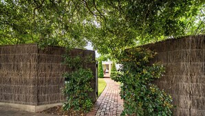 A brick pathway leading to a building with a thatched roof, surrounded by lush greenery and a tall, woven fence. - Geelong Road (Barwon Heads)