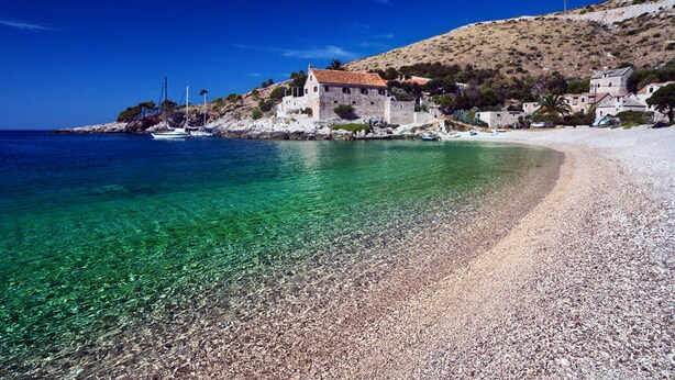 A coastal scene with clear turquoise waters, a pebble beach, and a stone building with a red-tiled roof. - Comfortable apartment in Stari Grad (Stari Grad)