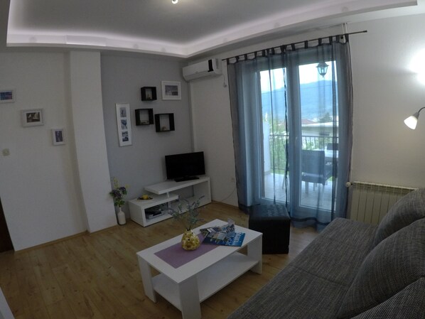 A living room with a white entertainment unit, a flat-screen TV, a coffee table with magazines, a grey sofa, and a balcony with a view. - Comfortable apartment in Stari Grad (Stari Grad)