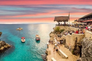 A coastal scene with boats on the water, a stone staircase leading down to the beach, and a crowd of people on the upper level of a building.