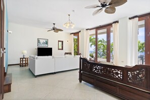 A spacious living room with a white sofa, a wooden bench, ceiling fans, and large windows offering a view of the outdoors.