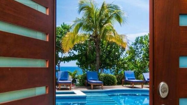 A wooden door leading to a swimming pool area with blue lounge chairs and palm trees. - Hospitality Expert - Little Waters Villa Negril (Negril)