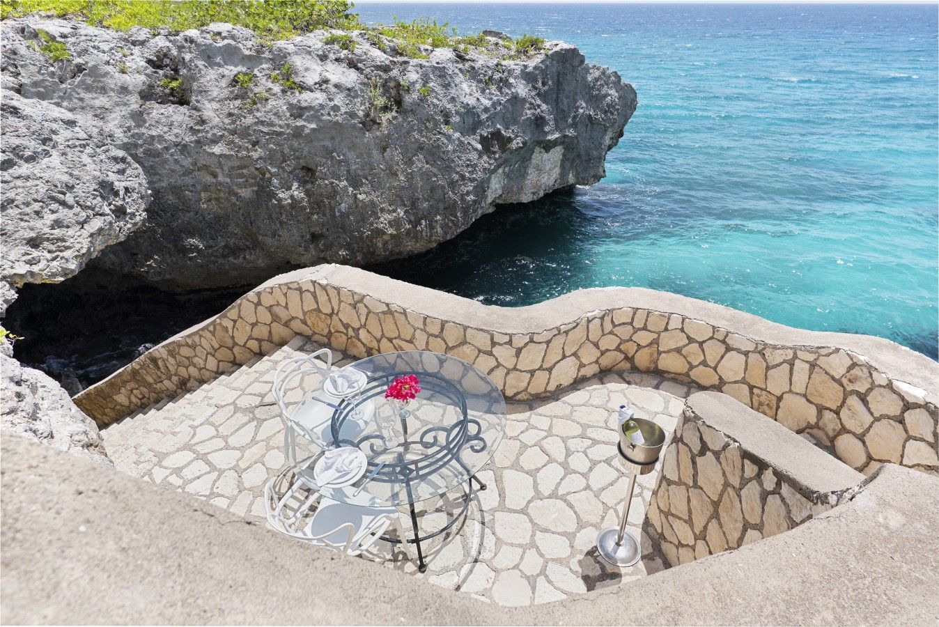 A stone-tiled outdoor seating area with a glass table and chairs, a flower, and a bottle of wine.