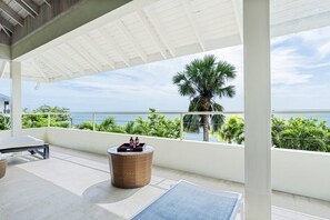 Ein überdachter Außenbereich mit Liegestuhl, einem kleinen runden Tisch mit Tablett und Blick auf das Meer und Palmen.