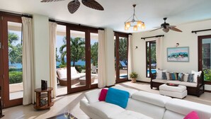 A spacious living room with a large white sofa, a coffee table, and a view of a pool and palm trees through glass doors. - Hospitality Expert - Little Waters Villa Negril (Negril)