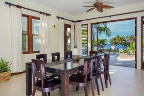 A dining area with a large table and chairs, a ceiling fan, and a view of the ocean through large windows.
