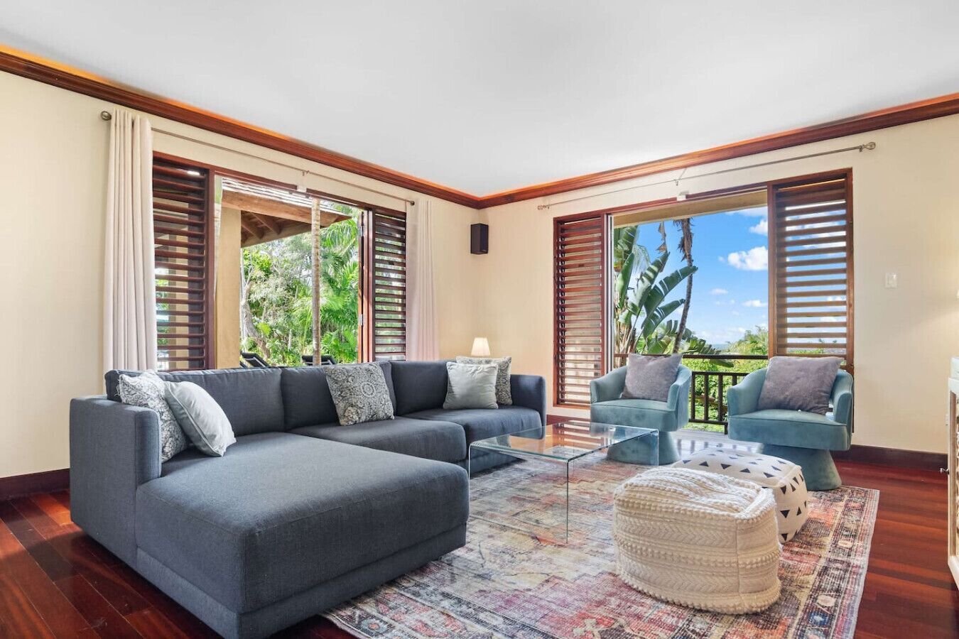 Un salón amplio con un sofá seccional gris, un sillón azul y una mesa de centro de vidrio. Ventanas grandes con contraventanas de madera ofrecen vistas a exuberante vegetación.