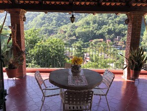 Un balcón con una mesa redonda y sillas, plantas en macetas y vistas a un paisaje verde exuberante con edificios a lo lejos.