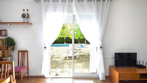 Une pièce dotée d’une porte-fenêtre coulissante donnant sur un jardin, d’une chaise en bois, d’un téléviseur posé sur un meuble en bois et d’une étagère ornée d’objets décoratifs.