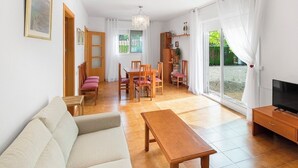 Un salon comprenant un canapé, une table basse en bois et un meuble pour la télévision. On y trouve également un coin repas avec une table et des chaises, ainsi qu’une grande fenêtre avec des rideaux.