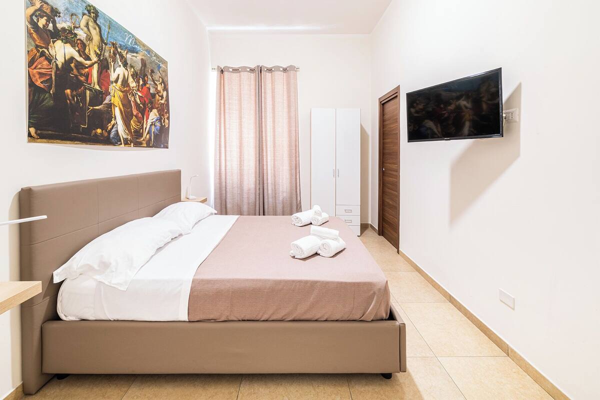A hotel room with a bed, a painting, a television, and a wardrobe.