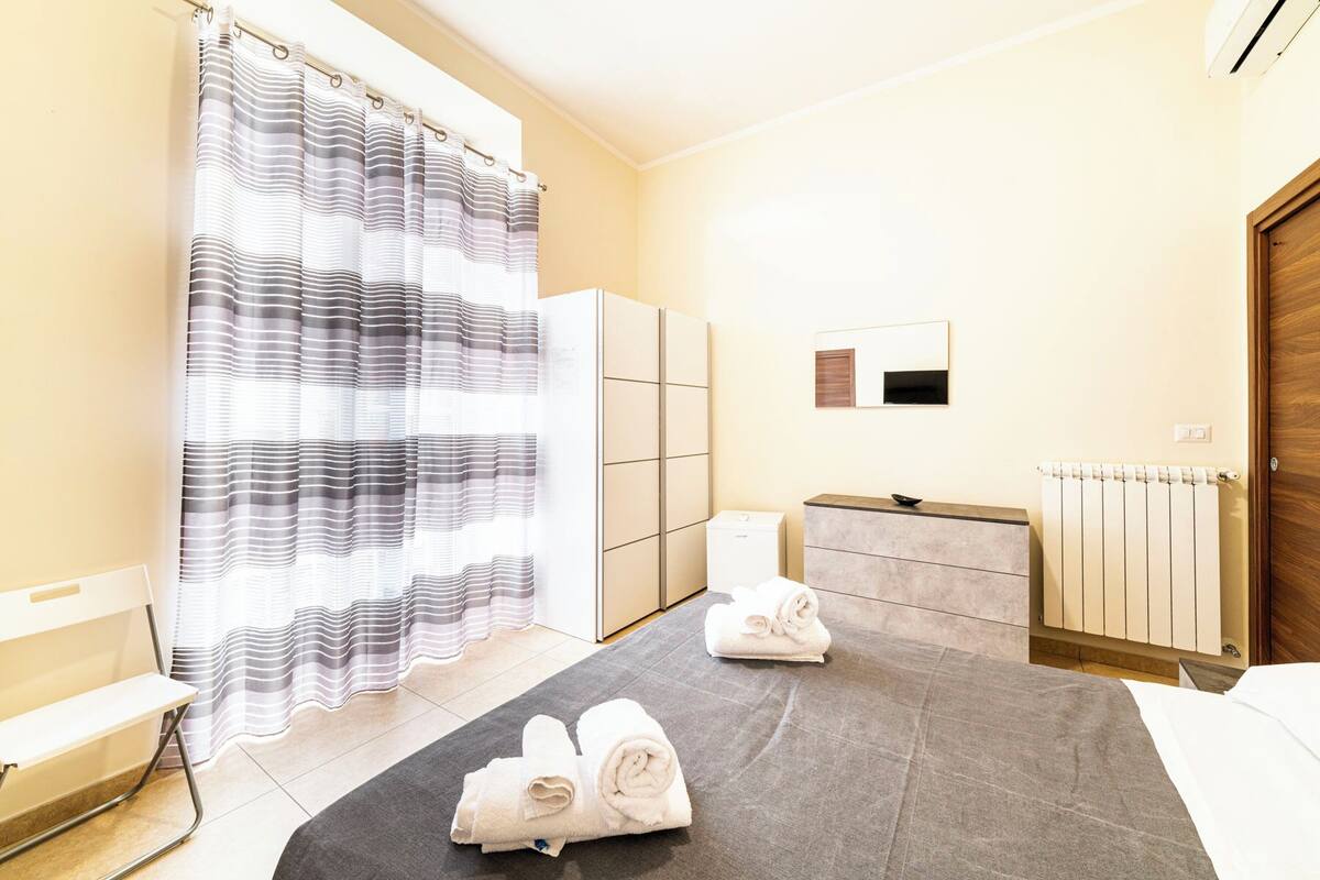 A hotel room with a shower curtain, a bed with a grey rug, two rolled towels, a wooden door, a radiator, and a small cabinet.