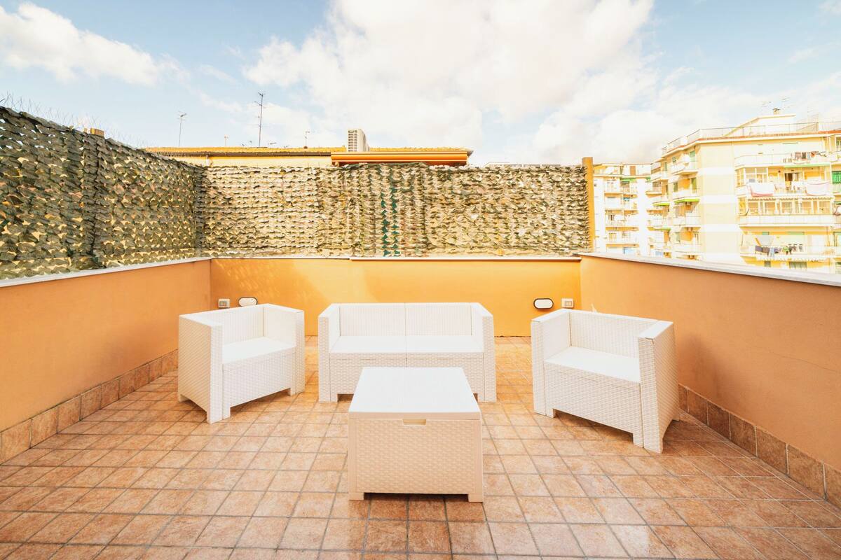 A rooftop terrace with wicker furniture, a stone wall, and a view of surrounding buildings.